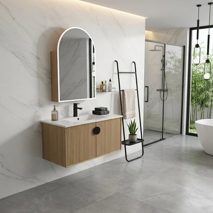 A modern wall-mounted bathroom vanity with a light wood ribbed cabinet, white countertop, black faucet, and an arched mirror with a warm white frame. The vanity is installed on a white marble-look wall in a contemporary bathroom with gray tile flooring, a glass shower enclosure, and a white freestanding bathtub.