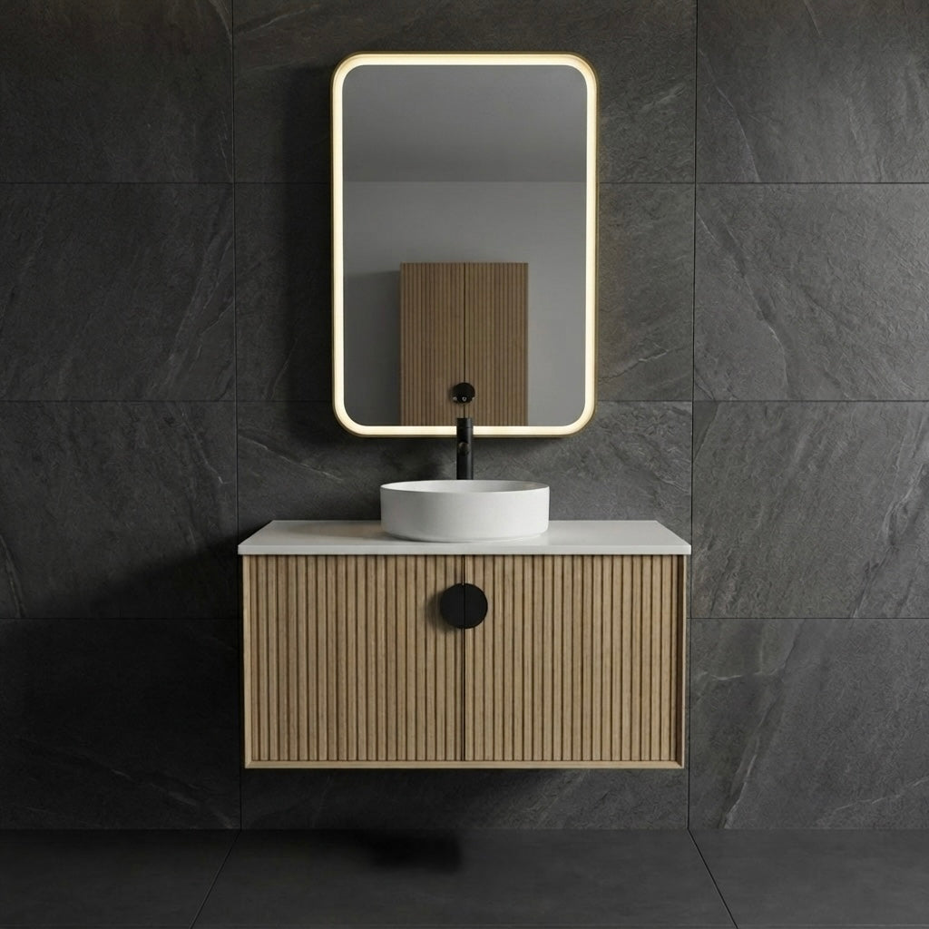 A modern wall-mounted bathroom vanity with a slatted wood cabinet, white countertop, white vessel sink, black faucet, and a rectangular backlit mirror, set against dark gray stone tile walls.