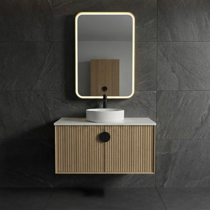 A modern wall-mounted bathroom vanity with a slatted wood cabinet, white countertop, white vessel sink, black faucet, and a rectangular backlit mirror, set against dark gray stone tile walls.