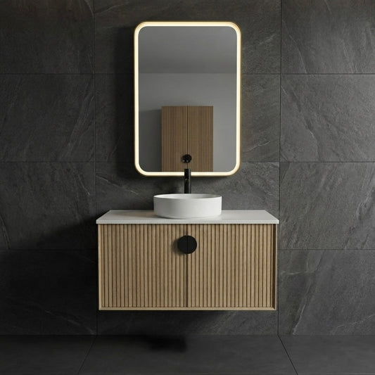 A modern wall-mounted bathroom vanity with a slatted wood cabinet, white countertop, white vessel sink, black faucet, and a rectangular backlit mirror, set against dark gray stone tile walls.