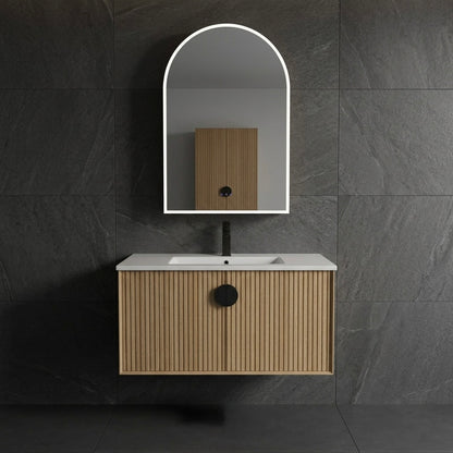 A modern wall-mounted bathroom vanity with a light wood slatted cabinet, white countertop, black faucet, and an arched LED-backlit mirror, set against a dark gray stone-tile wall.